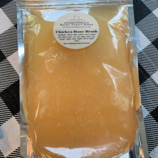 Golden color chicken bone broth is displayed on a checkered table cloth. The broth is contained in a sealed mylar bag.