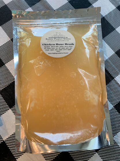 Golden color chicken bone broth is displayed on a checkered table cloth. The broth is contained in a sealed mylar bag.