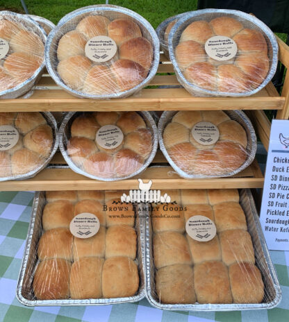 pans of golden brown soft sourdough dinner rolls on a shelf for purchase at an outdoor farmer's market