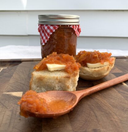 A small glass jar of deep orange colored marmalade sits behind a toasted roll and wooden spoon. Atop the toasted roll is a pat of butter and the orange marmalade.